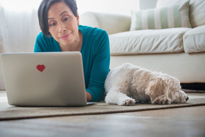 Older woman types on laptop on floor while dog rests next to her