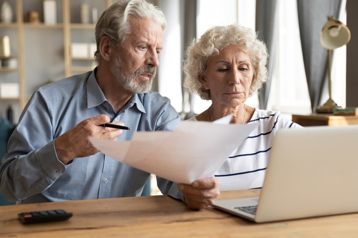 Older man holds documents and sits next to older woman at laptop