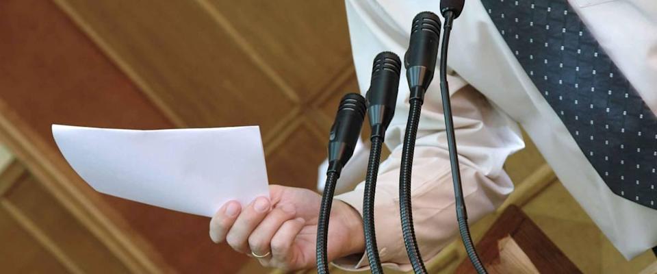 Man stands in front of several microphones, close up and talking while holding piece of paper.