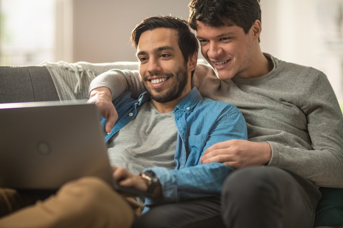 A couple on a couch is gazing at a laptop and smiling.